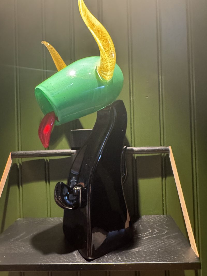 Glassfigur: grønt hode med gullhorn og rød tunge på en svart glasskropp, i en hylle