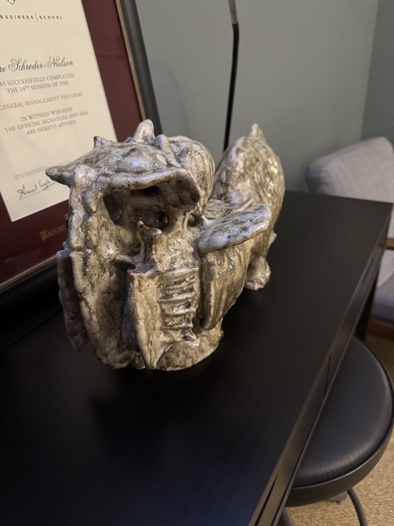 Keramikkskulptur av et fabeldyr med åpen kjeft, på et skrivebord, med et innrammet diplom bak