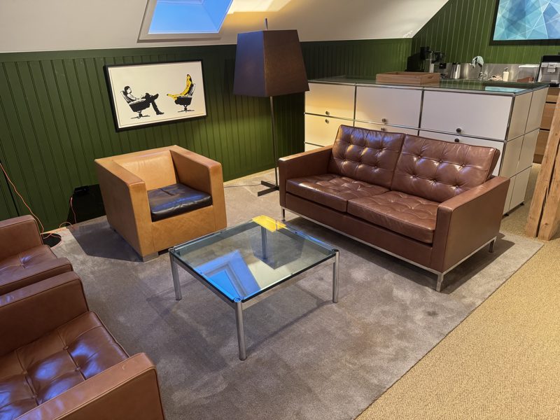 Lounge under skråtak: brun skinnsofa og to lenestoler rundt et glassbord, grønne panelvegger, kaffestasjon i bakgrunnen