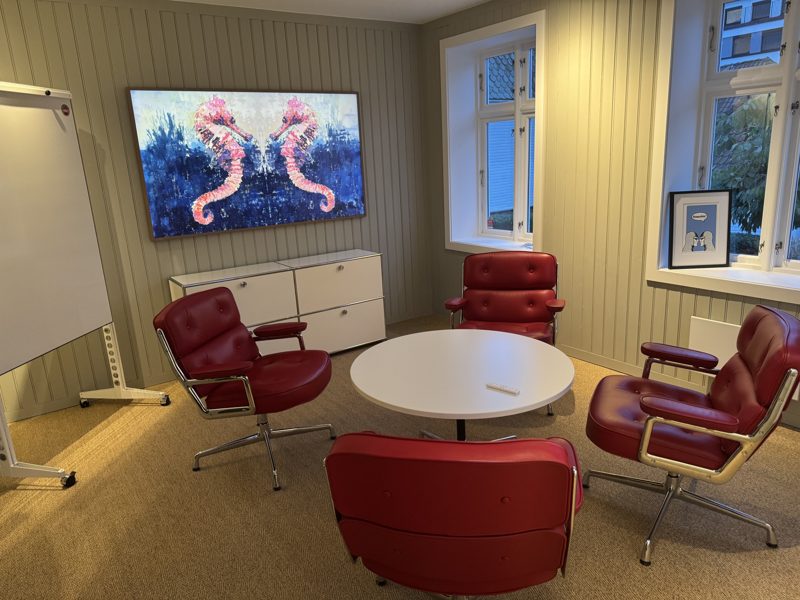 Møterom: rundt hvitt bord, fire røde skinnstoler, en stor skjerm på veggen som viser et kunstverk, flipover