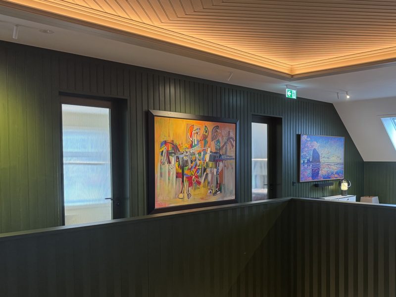Galleriet i andre etasje: et fargerikt maleri i svart ramme på grønn panelvegg, en glassdør ved siden av og gelenderet under