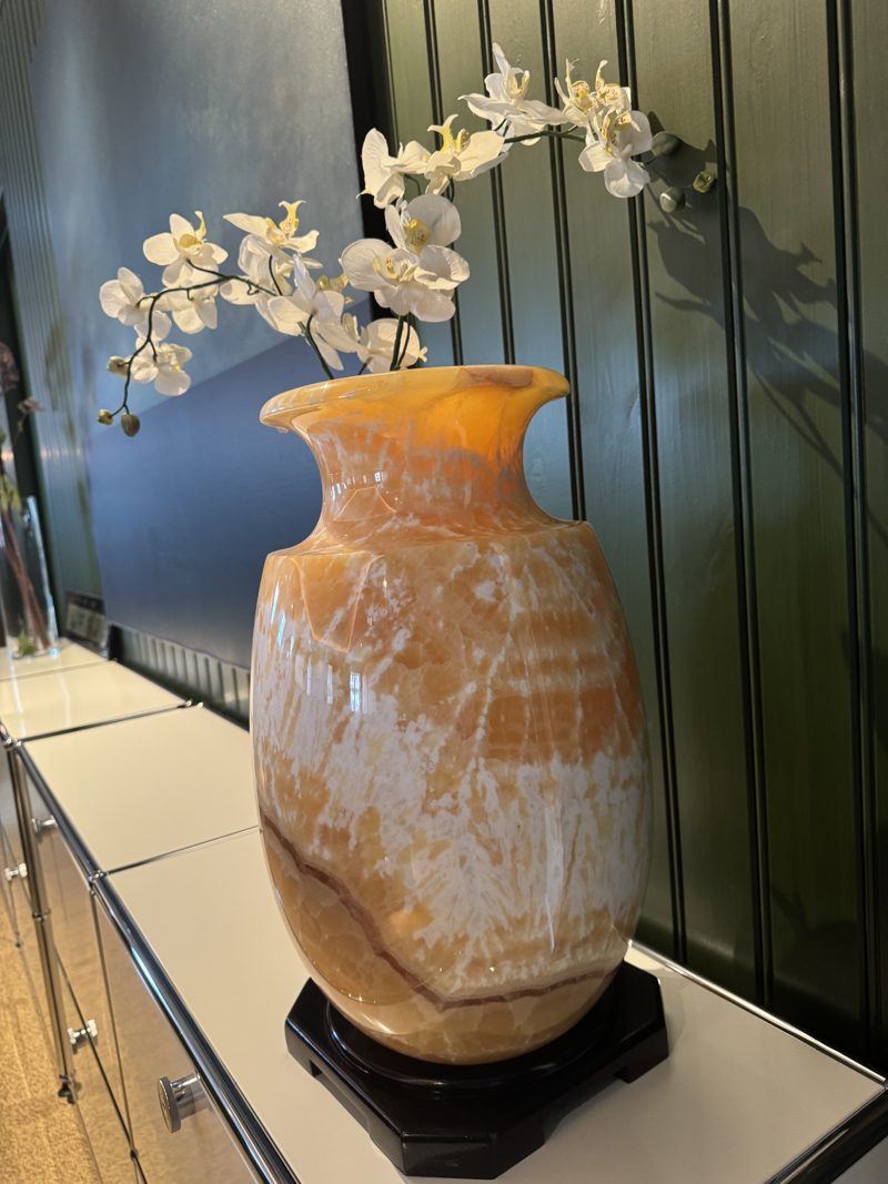 Høy vase i oransje og hvit stein på svart fot, med en hvit orkidé, mot grønn panelvegg